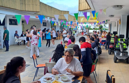 Imagem de mais de duas dezenas de pessoas em convívio. Algumas estão sentadas, à mesa, a conversar e a comer. Outras, no centro da imagem, estão a dançar.