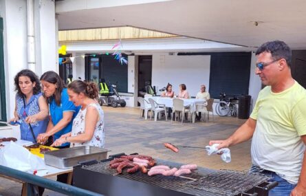 Em primeiro plano um churrasco com salsichas e chouriços em fase de preparação. Está uma pessoa a preparar os petiscos. Mais ao lado estão mais três pessoas a ajudar a cortar chouriços e salsichas já prontos. Ao fundo da imagem uma mesa, algumas cadeiras e três pessoas sentadas.