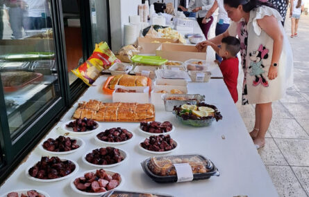 Aspeto de várias mesas com uma considerável diversidade de comida: saladas, frango no churrasco, chouriço, bolas e batatas fritas. Mais ao fundo muitos pratos e copos empilhados. Várias pessoas estão a servir-se (ou a avaliar o aspeto dos produtos oferecidos).