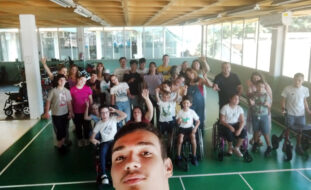 Selfie de grupo de pessoas em espaço interior do Centro de Reabilitação da APPC. Em primeiro plano rosto de quem fez a fotografia. Mais atrás perto de três dezenas de pessoas acenam para a fotografia.