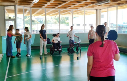 Oito jovens praticam Boccia. Em primeiro plano, de costas, uma pessoa assinala a cor da bola que ganha o ponto.
