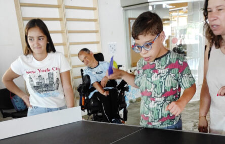 Três pessoas junto de uma mesa de Polybat. Mais atrás outra pessoa, em cadeira de rodas, assiste.