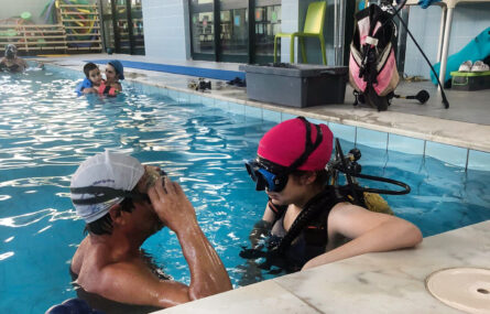 Perspetiva diagonal de piscina com cinco pessoas dentro de água. Em primeiro plano duas pessoas conversam. No chão, ao lado da piscina, está pousado algum material de mergulho.