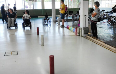 Várias pessoas em espaço desportivo interior. Duas, em cadeira de rodas, preparam-se para uma atividade. Várias outras assistem (ao fundo e na zona lateral).