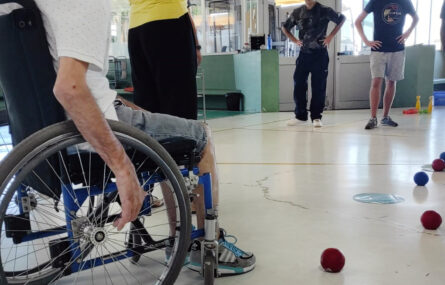 Pessoas a jogar Boccia em espaço interior. Algumas bolas espalhadas pelo chão. Em primeiro plano uma pessoa em cadeira de rodas está a olhar para quem faz a fotografia
