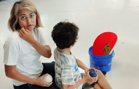 Pessoa adulta e criança no chão. Na mão seguram bolas de Boccia. Ao lado algum equipamento.
