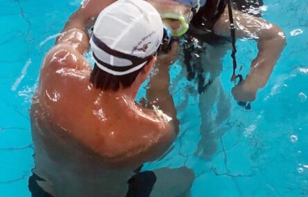 Duas pessoas dentro de uma piscina. De costas alguém ajuda outra pessoa (equipada com material de mergulho) a preparar algo.