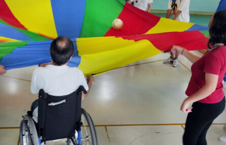 Várias pessoas, em círculo, levantam pano com várias cores em cima do qual está uma bola branca.