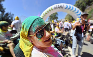 Fotografia de uma pessoa, sorridente, a usar óculos de sol. Está parcialmente coberta de pó colorido (de vários tons). Ao fundo, desfocadas, estão muitas mais pessoas.