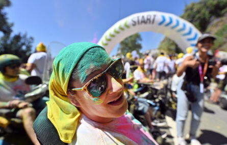 Fotografia de uma pessoa, sorridente, a usar óculos de sol. Está parcialmente coberta de pó colorido (de vários tons). Ao fundo, desfocadas, estão muitas mais pessoas.
