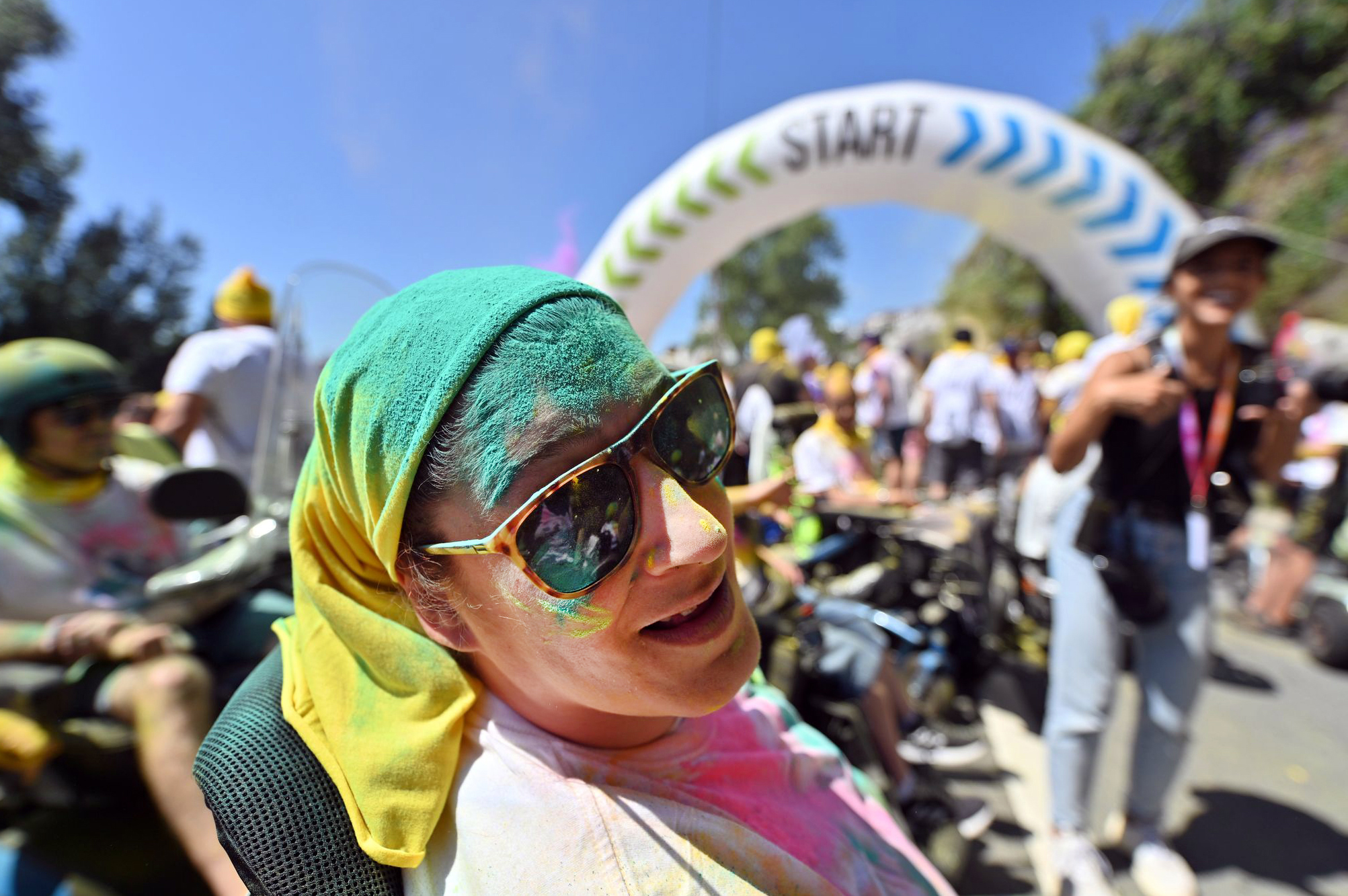 Fotografia de uma pessoa, sorridente, a usar óculos de sol. Está parcialmente coberta de pó colorido (de vários tons). Ao fundo, desfocadas, estão muitas mais pessoas.