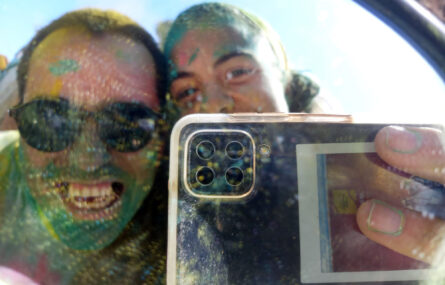 Duas pessoas fazem uma “selfie” com telemóvel e ao mesmo tempo fotografam-se em retrovisor de carro. Estão parcialmente cobertas de pó colorido (de vários tons).