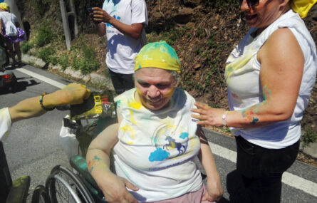 Pessoa sentada em cadeira de rodas sorri quando lhe espalham pó colorido em cima do braço.