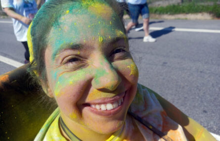 Grande plano de jovem a sorrir para a fotografia. Está parcialmente coberta de pó colorido (essencialmente amarelo e azul-esverdeado).