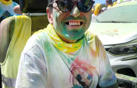 Pessoa sentada em cadeira de rodas sorri para a foto. Está coberta de pó colorido (azul e amarelo).
