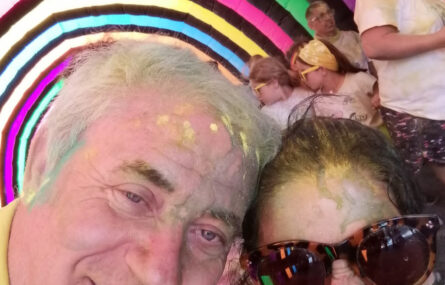 “Selfie” de duas pessoas sorridentes dentro de um túnel colorido. Estão parcialmente cobertas de pó azul e amarelo. Ao fundo ainda são visíveis mais participantes.