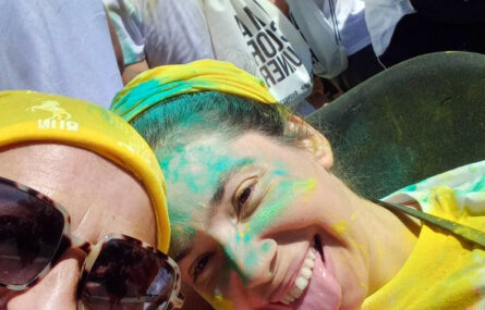 “Selfie” de duas pessoas sorridentes, sendo que uma delas está com a língua de fora. Estão parcialmente cobertas de pó azul e amarelo. Ao fundo ainda são visíveis mais participantes.
