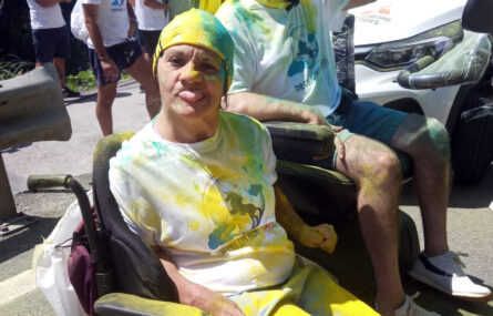 Fotografia de duas pessoas em cadeira de rodas. Estão parcialmente cobertas de pó azul e amarelo. Ao fundo está uma viatura de apoio, vendo-se ainda outros participantes