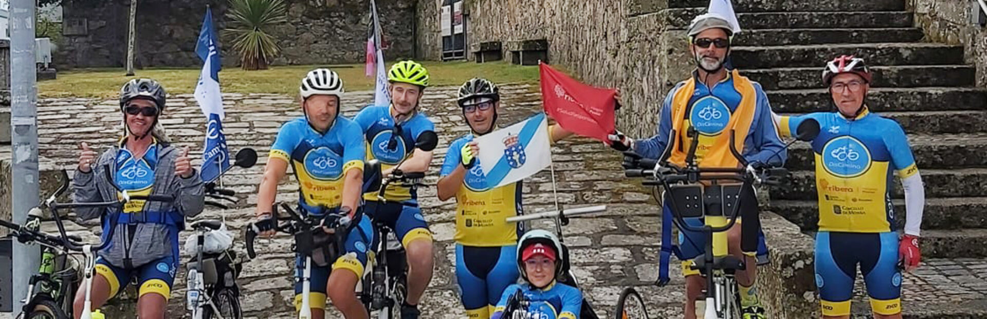 Grupo de sete pessoas em pé ou sentadas em bicicletas (normais e adaptadas). Ao fundo um edifício antigo.