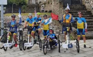 Grupo de sete pessoas em pé ou sentadas em bicicletas (normais e adaptadas). Ao fundo um edifício antigo.