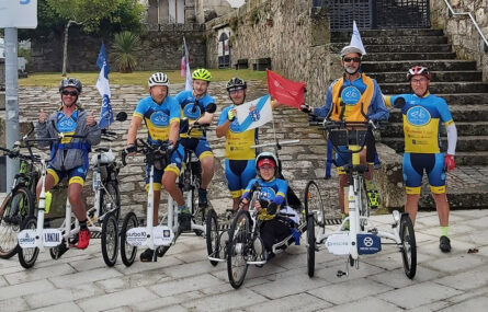 Grupo de sete pessoas em pé ou sentadas em bicicletas (normais e adaptadas). Ao fundo um edifício antigo.