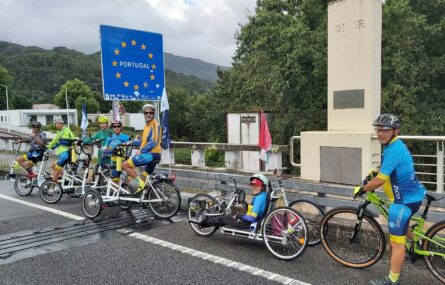 Grupo de sete pessoas bicicletas (normais e adaptadas) em estrada junto a uma placa que assinala a entrada (pela fronteira) em Portugal. Ao fundo árvores e montes.