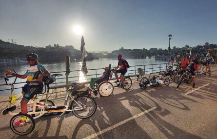 Grupo de 12 pessoas em bicicleta (normal ou adaptada) Fotografia junto do Rio Douro vendo-se do lado esquerdo parte da cidade de Gaia, à direita o Porto e, ao fundo, uma das pontes sobre o Rio Douro.
