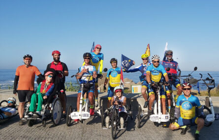 Grupo de 12 pessoas em pé ou sentadas em bicicletas (normais e adaptadas). Ao fundo vê-se o mar.