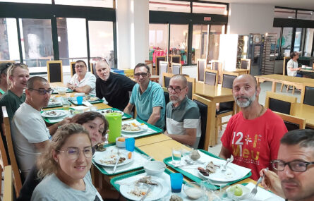 Fotografia de refeitório com várias pessoas sentadas à mesa durante uma refeição. Em cima da mesa estão pratos, talheres e copos. Mais ao fundo algumas mesas vazias. Ainda mais ao fundo outra pessoa também sentada à mesa.