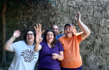 Quatro pessoas junto de um muro em pedra a abanar o braço direito e a dizer adeus para a fotografia.