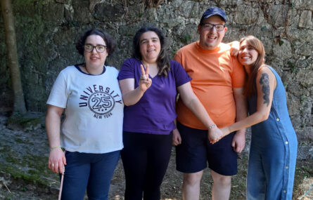 Quatro pessoas fotografadas junto a um muro em pedra. Uma das pessoas, com dois dedos da mão direita, faz o V de Vitória.