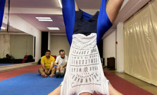 Pessoa a praticar “fly yoga”. Encontra-se “de pernas para o ar” pendurada pela cintura em pano azul suspenso do teto. Ao fundo outras duas pessoas assistem.