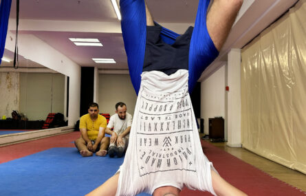 Pessoa a praticar “fly yoga”. Encontra-se “de pernas para o ar” pendurada pela cintura em pano azul suspenso do teto. Ao fundo outras duas pessoas assistem.