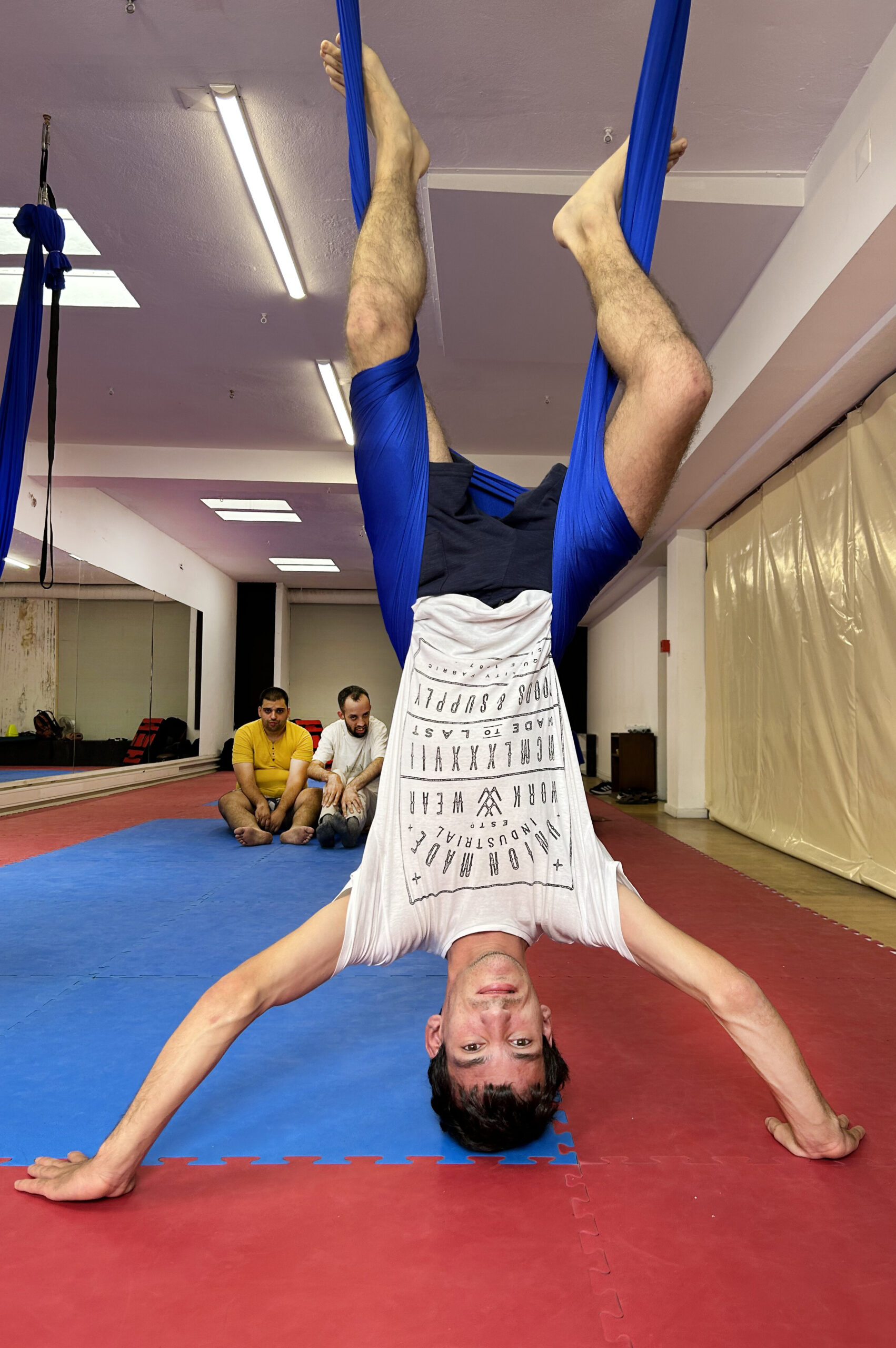 Pessoa a praticar “fly yoga”. Encontra-se “de pernas para o ar” pendurada pela cintura em pano azul suspenso do teto. Ao fundo outras duas pessoas assistem.