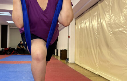 Pessoa a praticar “fly yoga” pendurada pela cintura em pano azul suspenso do teto.