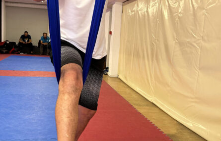 Pessoa a praticar “fly yoga” pendurada pela cintura em pano azul suspenso do teto.
