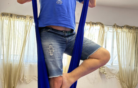 Pessoa a praticar “fly yoga” em pano azul suspenso do teto.