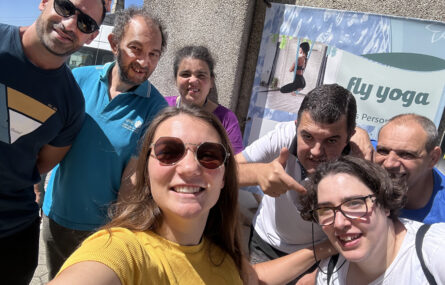 Grupo de sete pessoas a fazerem uma “selfie”. Mais ao fundo, pendurado em parede exterior de edifício, está um cartaz de promoção do “fly yoga”.