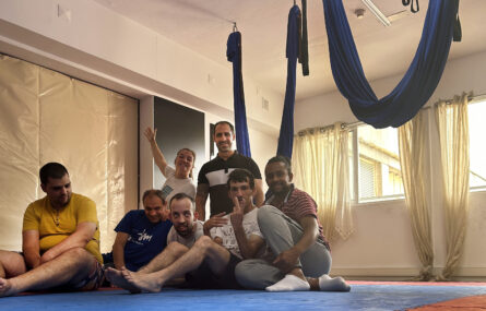 Sete pessoas sentadas ou de joelhos no chão. No teto estão suspensos dois panos azuis para a prática do “fly yoga”.