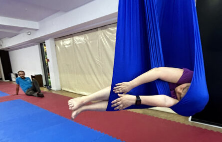 Pessoa deitada e suspensa em pano azul de prática de “fly yoga”. Ao fundo, sentada no chão, está outra pessoa a assistir.
