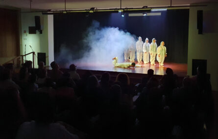 Fotografia interior do auditório da Junta de Freguesia de Paranhos. Em primeiro plano, na escuridão, pessoas sentadas. Em palco alguns atores e atrizes. Vê-se uma nuvem de fumo a encobrir parte dos atores.