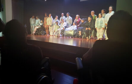 Atores e atrizes alinhados em palco durante os agradecimentos finais. Em primeiro plano, na escuridão, vê-se o recorte de duas pessoas sentadas em cadeiras de rodas.
