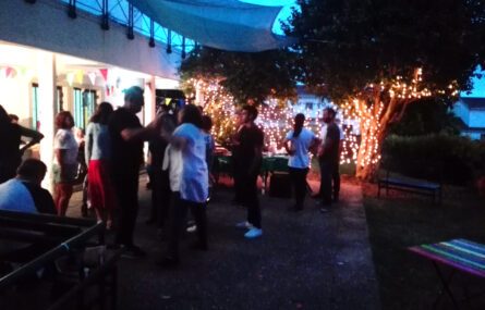Foto noturna de espaço exterior. Ao fundo fileiras de luzes iluminam o espaço. Algumas pessoas aparentam dançar.