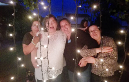Quatro pessoas, abraçadas, fazem retrato sendo que à frente delas estão penduradas algumas fileiras com pequenas luzes.