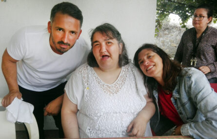 Três pessoas sorriem para a foto. Mais atrás surge outra pessoa.
