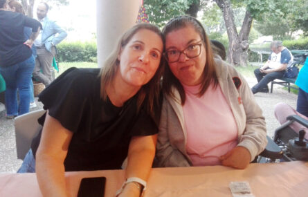 Duas pessoas sorriem para a foto sentadas junto de uma mesa. Ao fundo árvores e um jardim (com mais algumas pessoas na conversa).