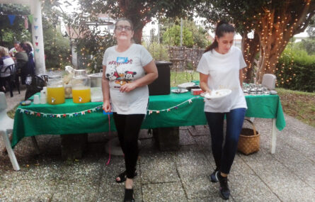 Duas pessoas aparentemente a dançar. Atrás delas uma mesa com pano verde e inúmeros pratos, copos, taças e utensílios lá pousados. Ao fundo da imagem árvores e jardim. Nas árvores estão penduradas fileiras com pequenas luzes.