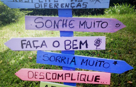 Placa de sinalização colocada em relva de jardim com sete setas de distintas cores. Cada uma das setas apresenta uma mensagem. Seja gentil. Respeite as diferenças. Sonhe muito. Faça o bem. Sorria muito. Descomplique. Agradeça!