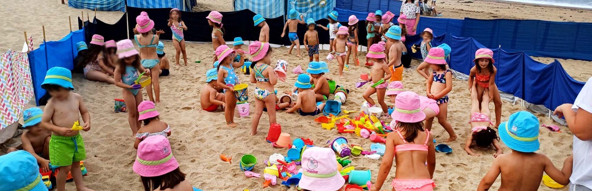 Fotografia de cerca de quatro dezenas de crianças na praia. A areia está repleta de brinquedos. Ao fundo estão seis barracas de praia. Vê-se o mar e, mais longe ainda, a Capela do Senhor da Pedra (junto ao mar). Todos os rostos das crianças foram desfocados.