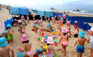 Fotografia de cerca de quatro dezenas de crianças na praia. A areia está repleta de brinquedos. Ao fundo estão seis barracas de praia. Vê-se o mar e, mais longe ainda, a Capela do Senhor da Pedra (junto ao mar). Todos os rostos das crianças foram desfocados.